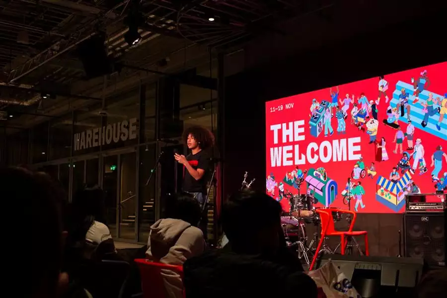 A group of children watching a person speak on stage, with the words 'The Welcome' on a screen behind them