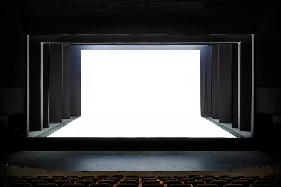 A theater stage with a dramatic, modern design is shown, featuring black framing and a stark white, illuminated backdrop. The contrast between the dark seating area and the bright stage creates a striking visual effect.