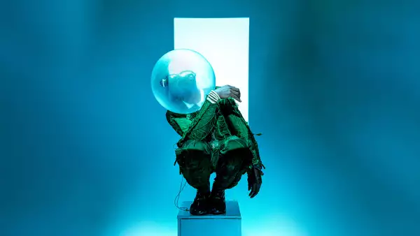 Benji Reid crouched on a small pedastal in front of a white strip against a blue background. He is wearing a green pattered suit and a bubble sculpture on his head.