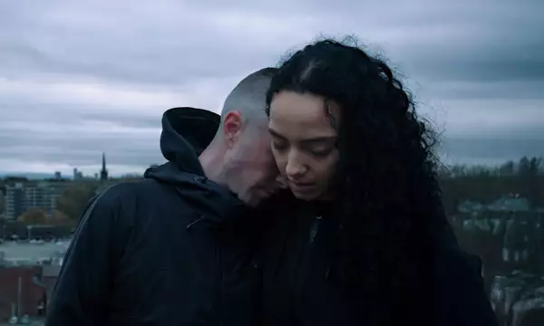 A man in a black hooded jacket leans his head against a woman’s shoulder, his eyes closed, while she looks downward with a solemn expression. The background shows an overcast sky and cityscape.