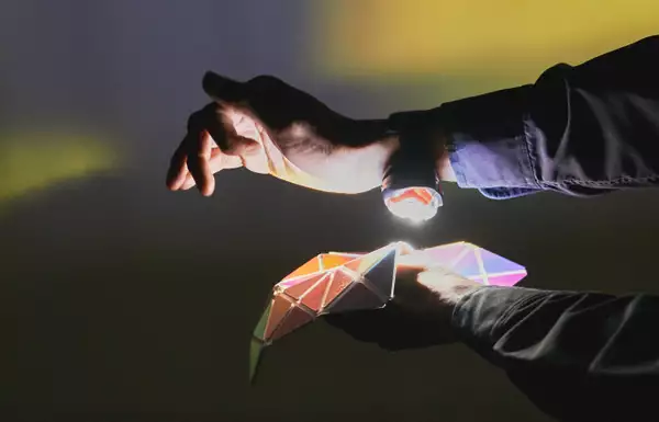 In a dark room, two arms come into shot from the right. The top arm has a small bright light strapped to the underside of the wrist, which is pointing down towards a kaleidoscopic, geometric matrix of mirror shapes held up by the other hand. Yellow and green coloured light reflects off the mirrored shapes onto the wall behind.