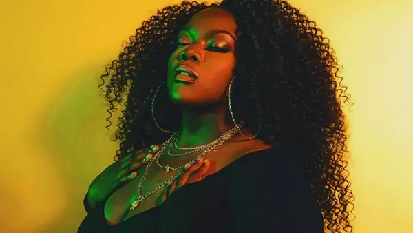 Dija Bhule looking up with eyes close bathed in blue light and against a.yellow backdrop