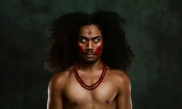 A member of FAFSWAG with glowing white eyes and red and black face paint stares intensely at the camera. They wear red beaded necklaces. The background is dark green.