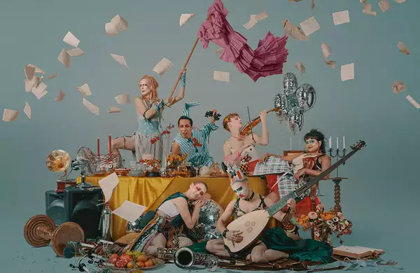 A group of performers around a table littered with objects, some are playing musical instruments, one is waving a flag