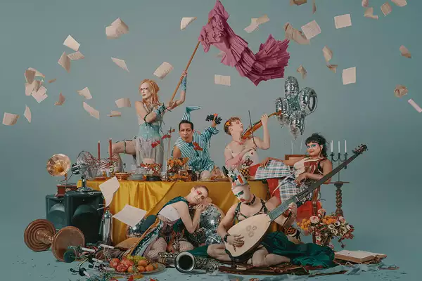 A group of performers around a table littered with objects, some are playing musical instruments, one is waving a flag