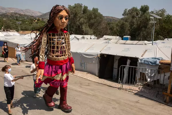 The Little Amal puppet walks past refugee camps in Greece. The puppeteers are wearing face masks.