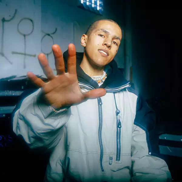 The artist is33n putting his hand up towards the camera. He is wearing a silver metallic coat.