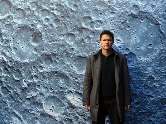 Luke Jerram, a male artist stood in front of a close up of a moon crater artwork