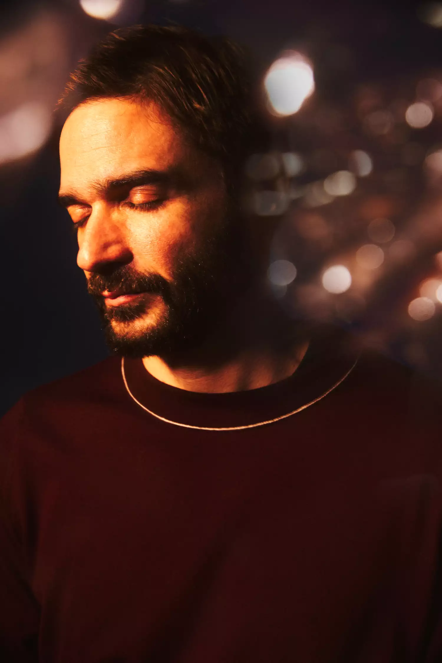 Headshot of Jon Hopkins with his eyes closed. He is wearing a maroon jumper and gold chain. The background shows blurry lights.