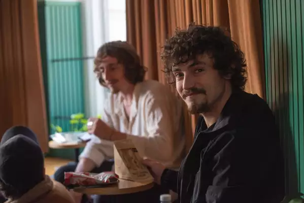 Three young men are sat eating sandwiches with a curtain behind them. One is looking at the camera while the other two are out of focus.