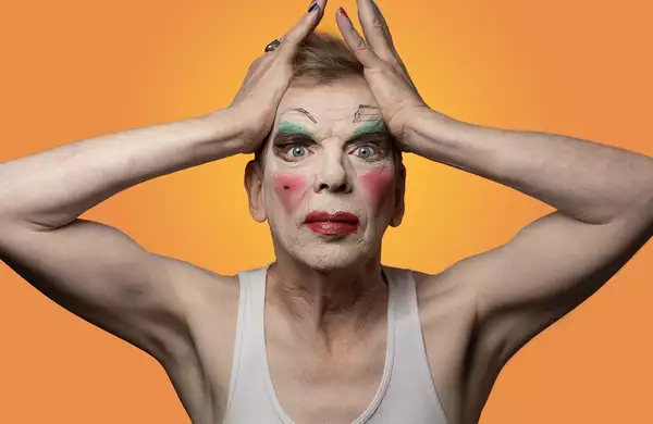 David Hoyle bold makeup and hands either side of his forehead against an orange background