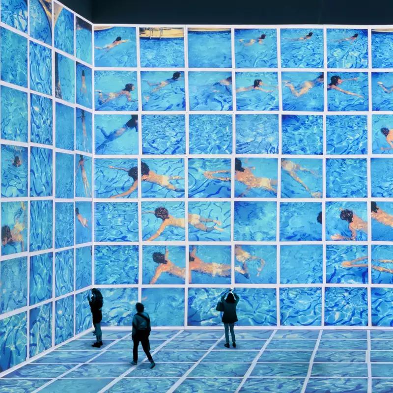 Three people stood in the David Hockney exhibition at Lightroom. His swimming pool paintings are shown on the screens.