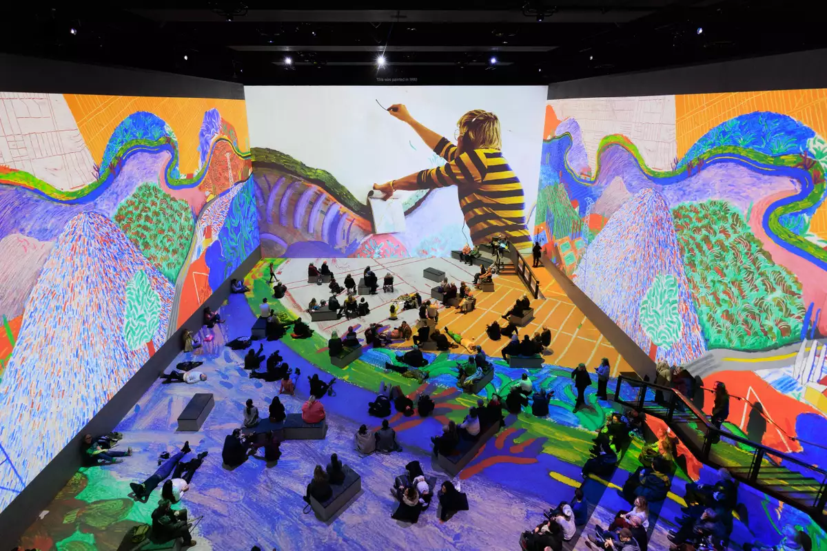 Photograph of David Hockney: Bigger & Closer (not smaller & further away) at Lightroom taken from above. It shows people sitting and lying on the floor, immersed inside Hockney's artworks.