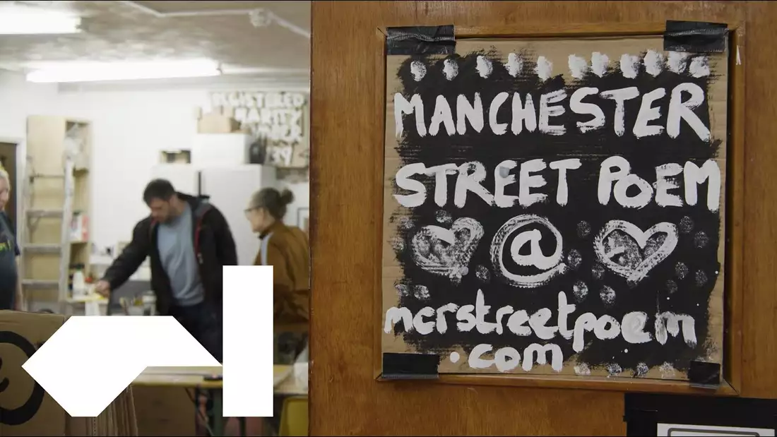 A sign saying Manchester Street Poem in the foreground with three people in the background working in a studio space