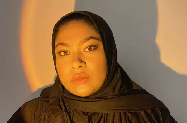 A headshot of a woman looking straight to camera. She wears a black top and scarf which partially covers her hair.