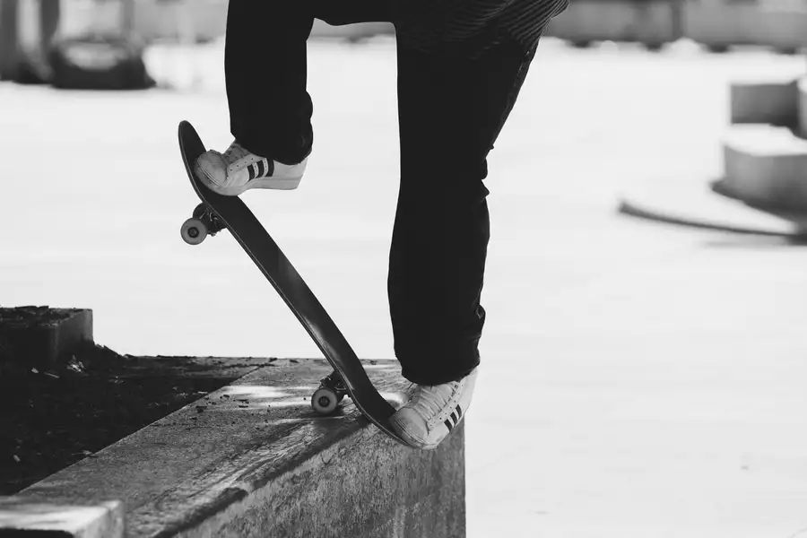 Black and white photograph of two feet on a skateboard. They are wearing white adidas trainers.