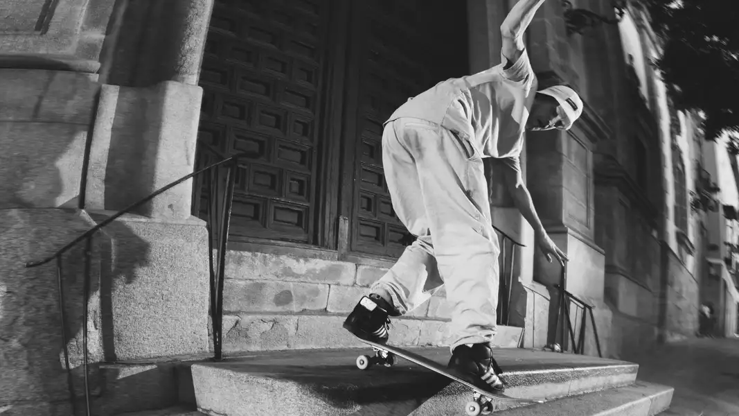 Black and white photograph of a skateboarder doing a trick on the steps of an old building, and wearing adidas trainers