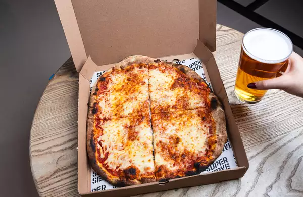 An open pizza box on a wooden table with a cheese pizza cut into four slices. A hand with red-painted nails holds a pint of beer beside it.