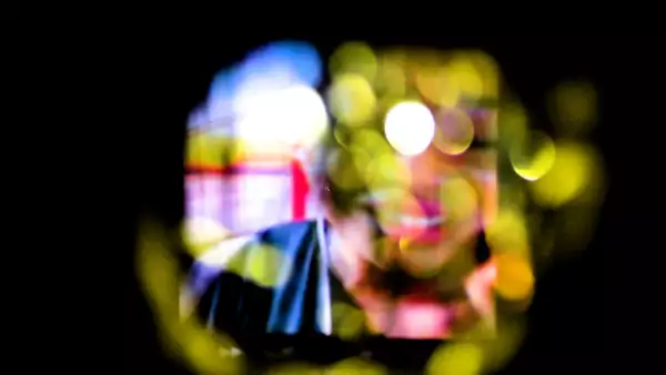 A blurred and light-distorted image down a dark camera lens of a smiling person who has black skin and is wearing pink lipstick.