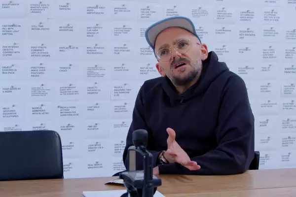 The artist Ryan Gander in his studio. He is a white man, wearing a black hoody and trousers and a multi-coloured cap.