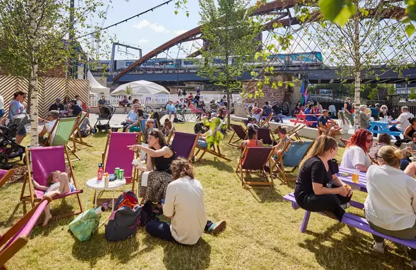 Factory Square filled with people sat on the grass and deckchairs, drinking and chatting. The sun is shining.