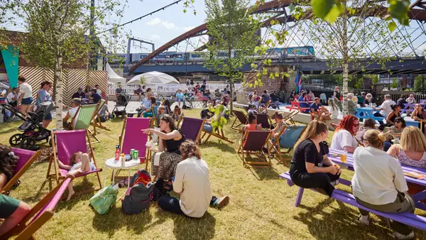 Factory Square filled with people sat on the grass and deckchairs, drinking and chatting. The sun is shining.