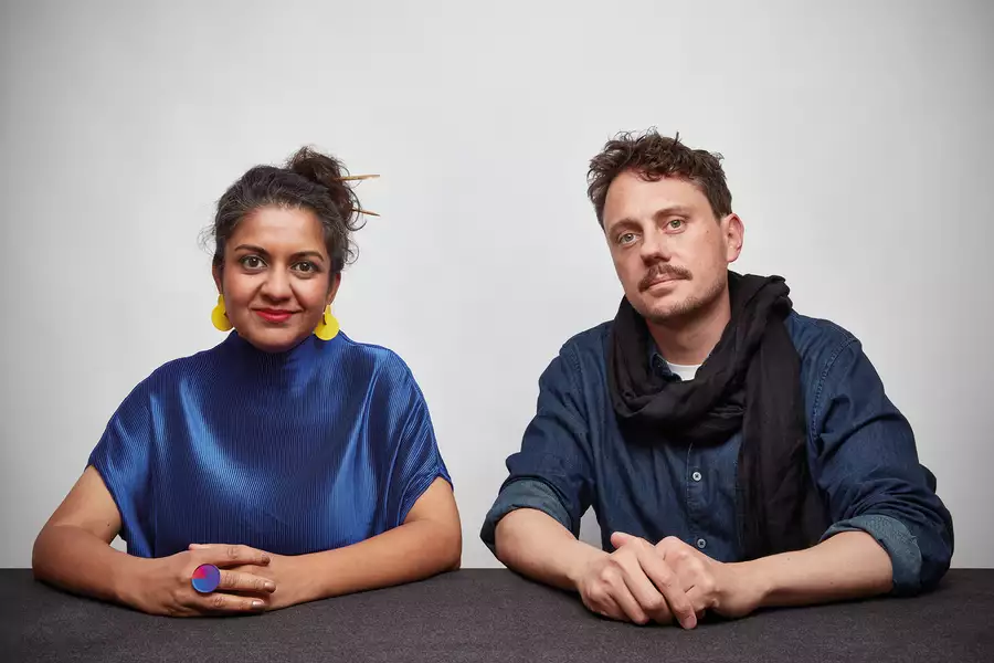 Two members of Superflux sat at a table, looking at the camera. The person on the left is wearing a shiny blue top and yellow earrings. The person on the right is wearing a blue shirt and black scarf.