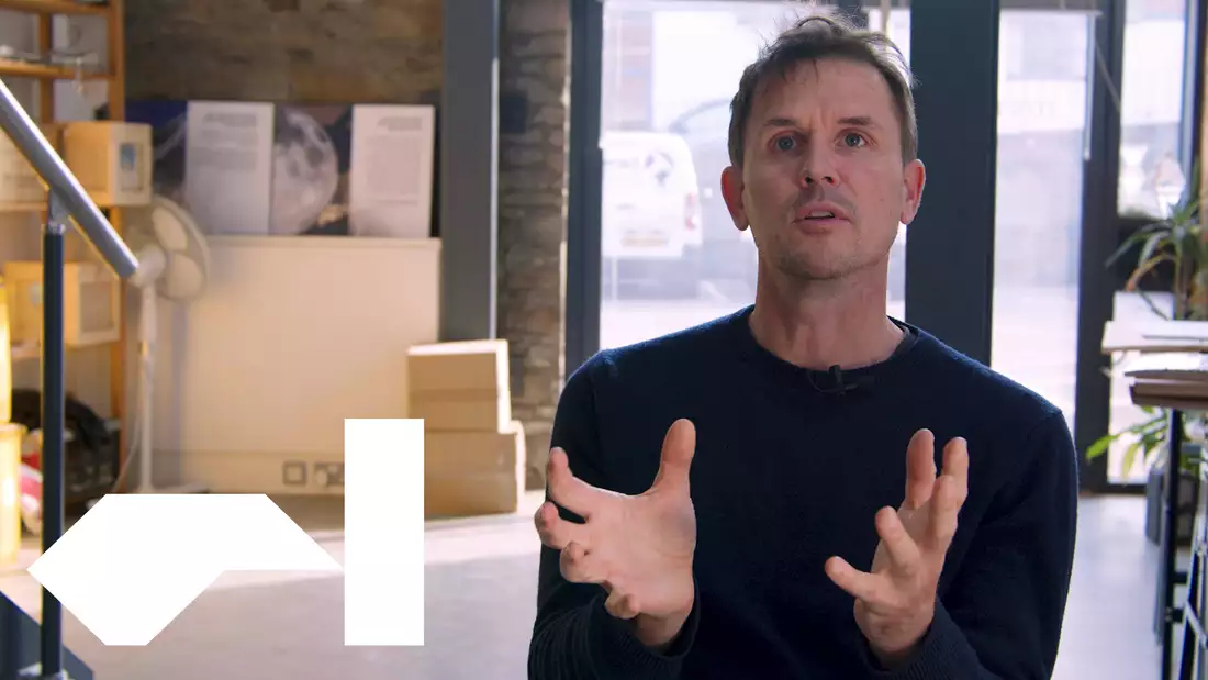 The artist Luke Jerram sits in his studio with boxes, natural light and plants visible behind him.