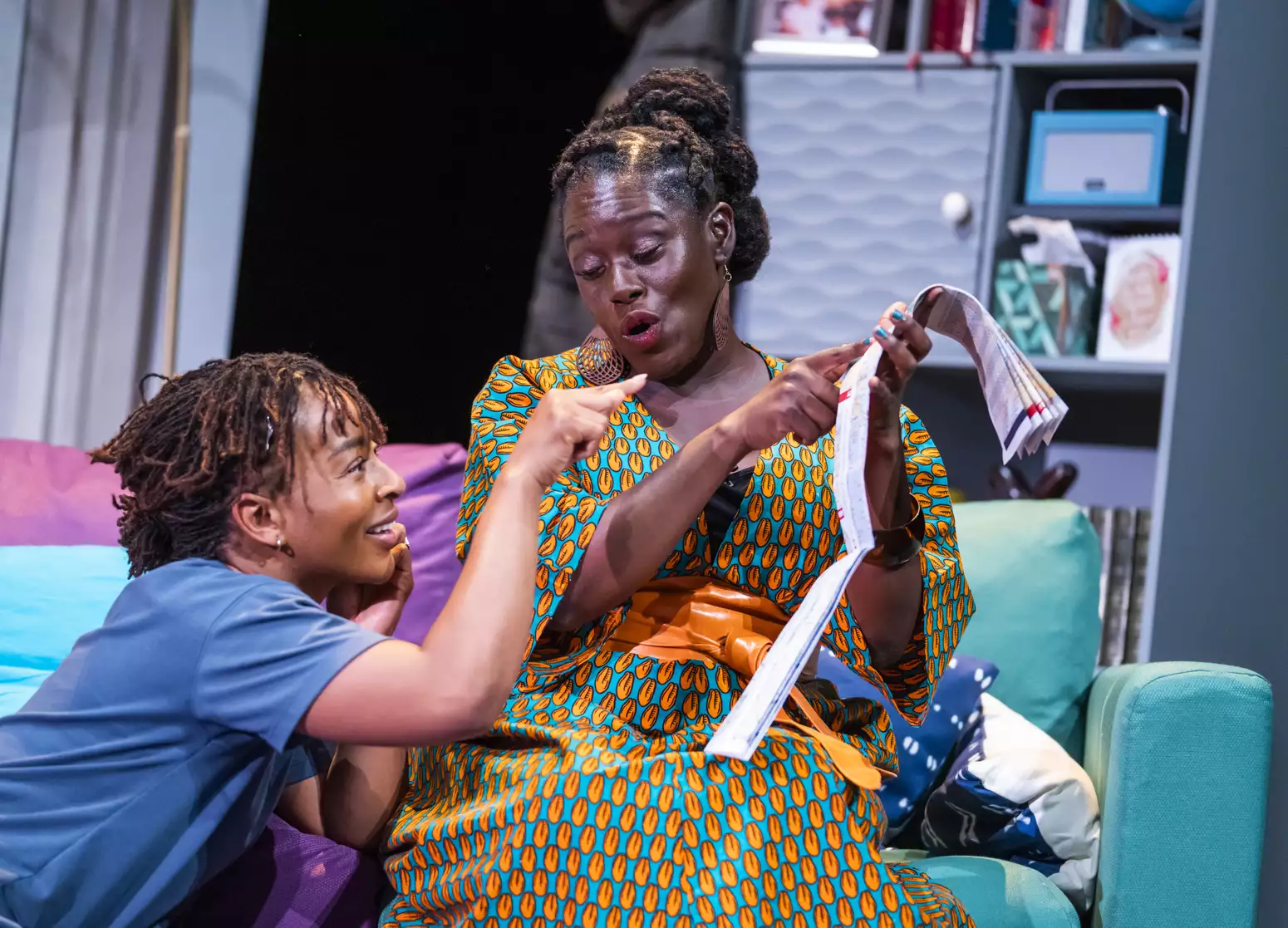 Uche Abuah and Michelle Asante on stage in Notes on Grief. Asante sits on a blue and purple sofa and holds up a folded paper map, which she points to. Abuah sits at her feet, smiling and also points at the map. Asante is Black with dark brown skin; her hair is twisted from the crown into three buns at the back of her head; she wears a bright orange and turquoise patterned dress and an orange wrap belt. Abuah is Black with medium brown skin; her hair is in fine locks, pulled back; she wears a blue T-shirt.