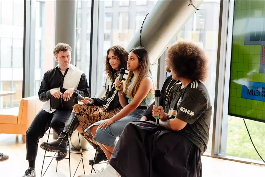 Four people sat on high chairs in front of a large window during a panel talk. One is speaking. There is a screen behind them with Dazed and Mubi branding on.