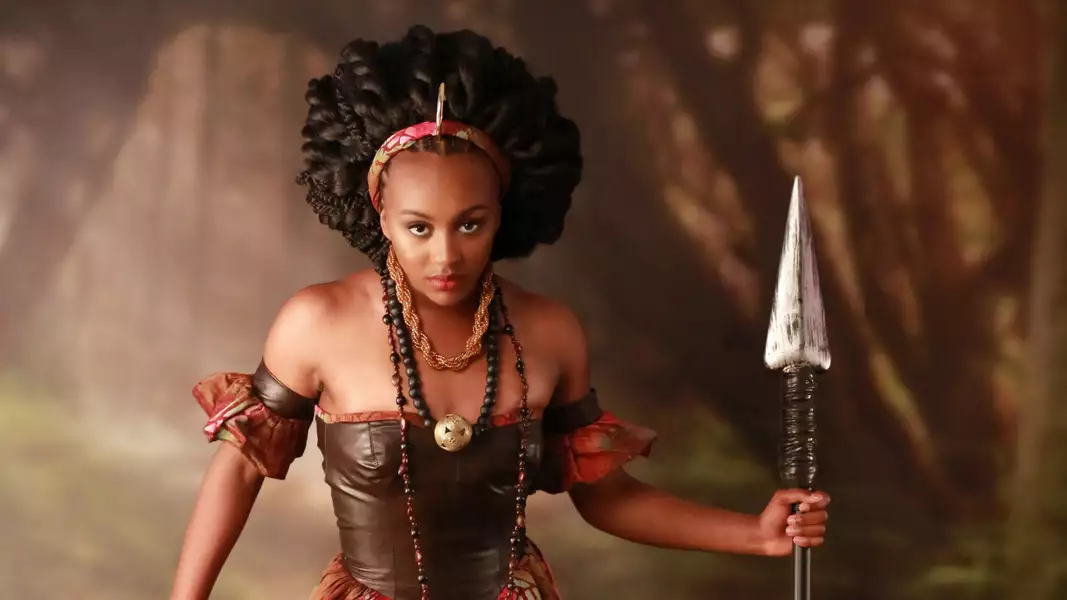 A woman dressed like a warrior in a leather top, holding a spear with a determined expression. Her hair styled is in twists with a headband. The background is blurred.