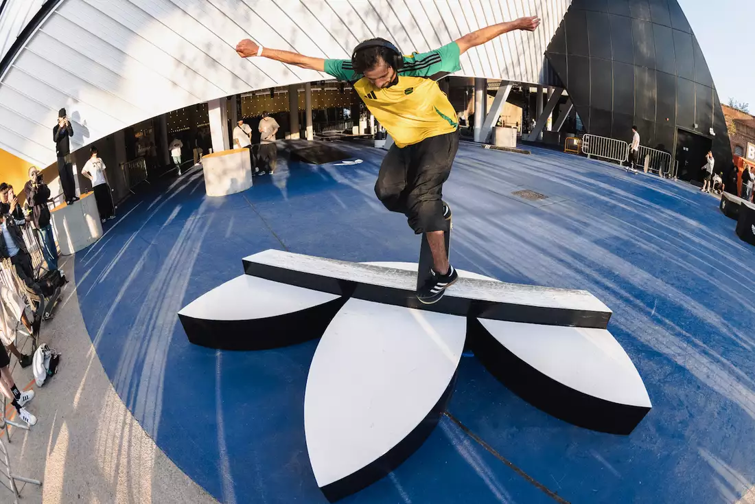 A skateboarder doing a trick on a ramp, which is the shape of the Adidas logo. There is a crowd of people watching outside Aviva Studios.