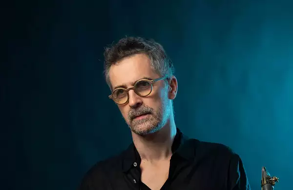 Sylvain Rifflet wearing round glasses stood on a deep blue background. The top of his saxophone reed is visible in the bottom right hand corner.