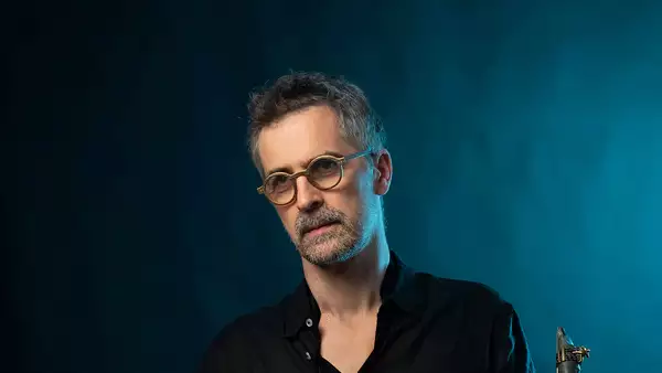 Sylvain Rifflet wearing round glasses stood on a deep blue background. The top of his saxophone reed is visible in the bottom right hand corner.
