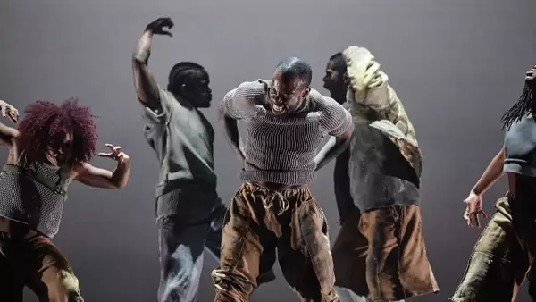 Five contemporary dancers perform with intense, expressive movements on stage, dressed in earthy-toned clothing. The central dancer leads the powerful scene, surrounded by others in dynamic, dramatic poses.