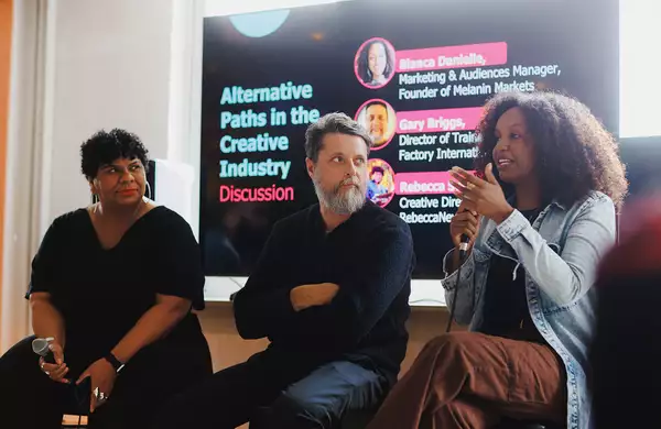 A panel discussion on "Alternative Paths in the Creative Industry" features three speakers seated in front of a large screen displaying their names and roles. One speaker is actively speaking into a microphone while the others listen attentively.