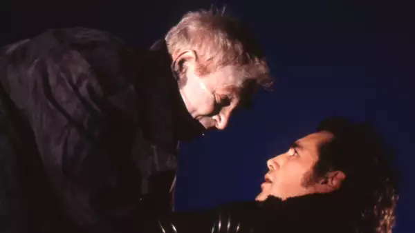 Russell Enoch towering over Mark Rylance, who is looking up at him in fear. Russell has his hands on Mark's neck. Both are wearing black coats.