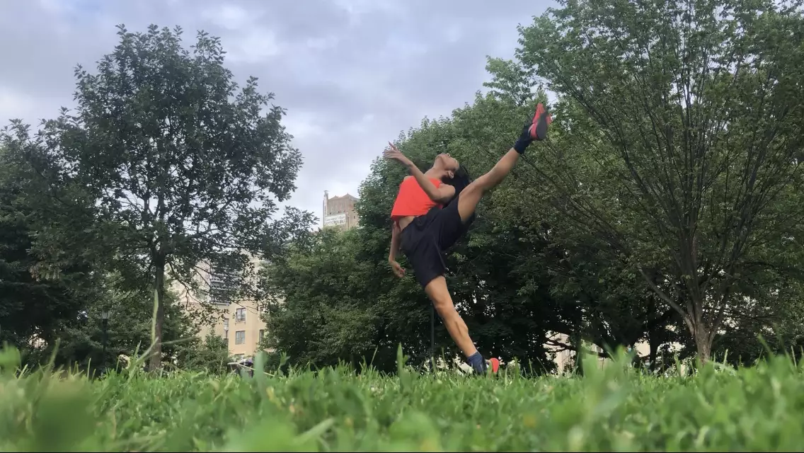 A horizontal photo of Benedict Nguyễn, wearing black shorts, orange tank, and black and orange sneakers, standing on right leg with left leg raised to the side, left arm bent in front of her torso, head tipped back, in a field with green grass and trees, with buildings in distant background.