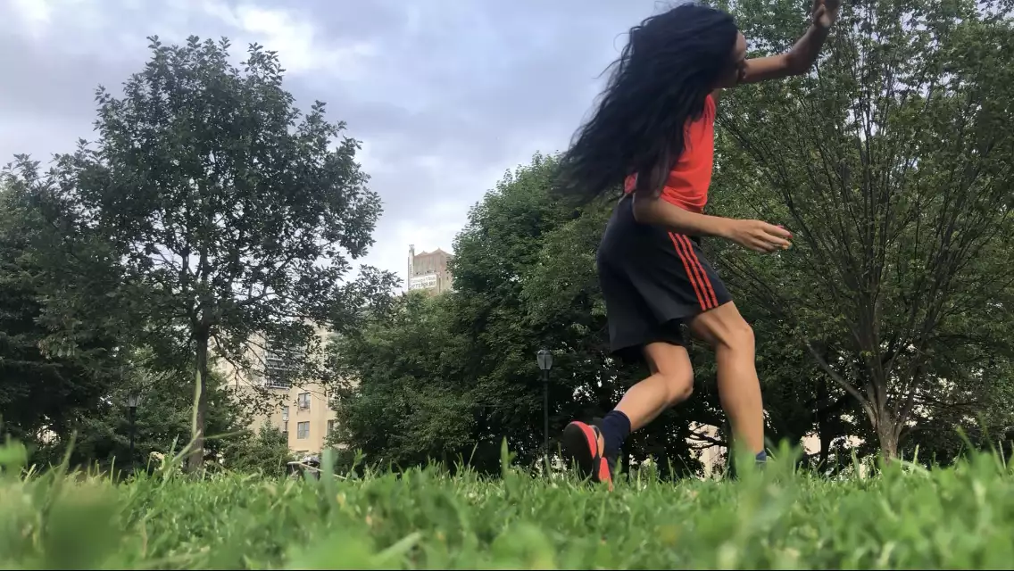 A horizontal photo of Benedict Nguyễn, wearing black shorts, orange tank, and black and orange sneakers, lunging forward on right leg with left arm reaching across her face, in a field with green grass and trees, with buildings in distant background.