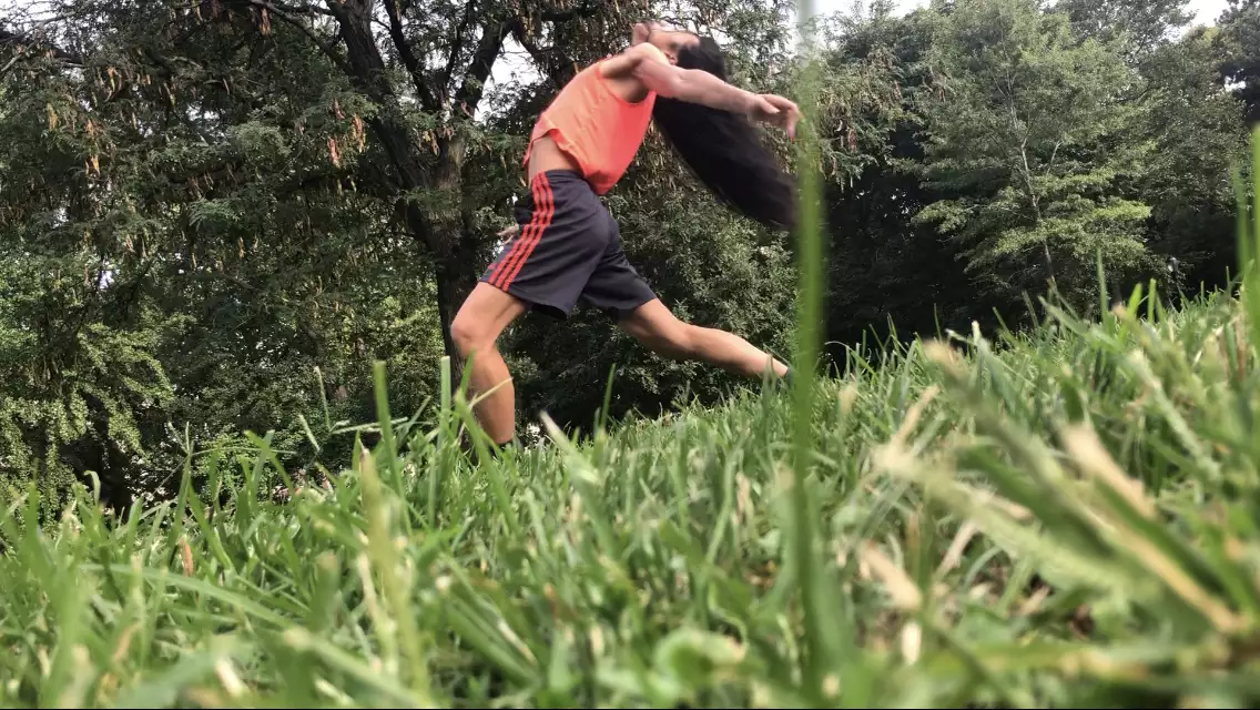 A diagonal photo of Benedict Nguyễn, wearing black shorts, orange tank, and black and orange sneakers, lunging forward on left leg with back arched left arm reaching behind her body, in a field with green grass and trees in background.