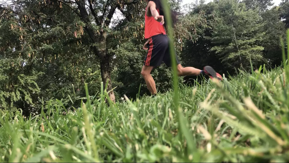 A diagonal photo of Benedict Nguyễn, wearing black shorts, orange tank, and black and orange sneakers, lunging forward on left leg facing away from camera with left arm reaching behind her body, in a field with green grass and trees in background.