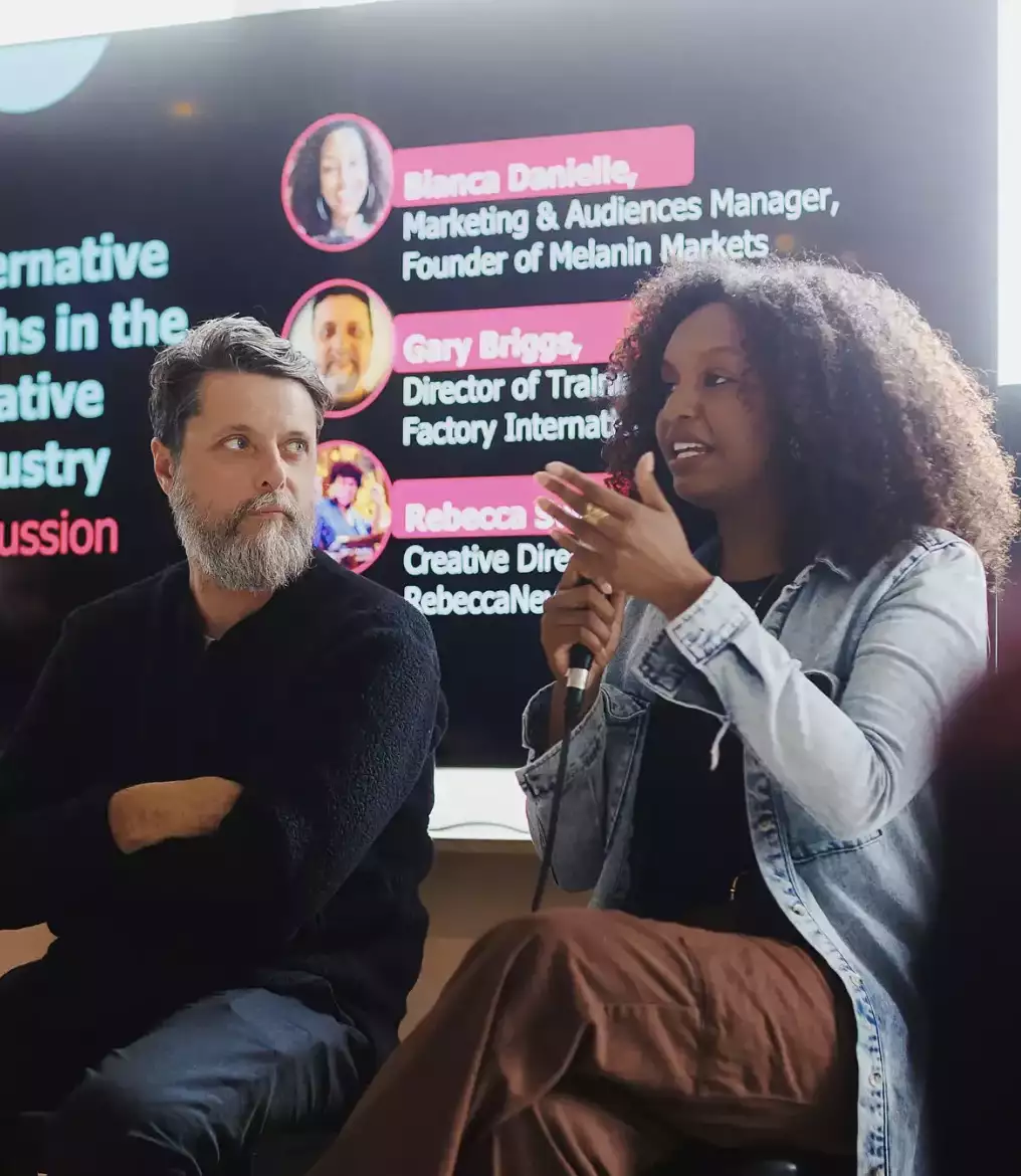 A panel discussion on "Alternative Paths in the Creative Industry" features two speakers seated in front of a large screen displaying their names and roles. One speaker is actively speaking into a microphone while the others listen attentively.
