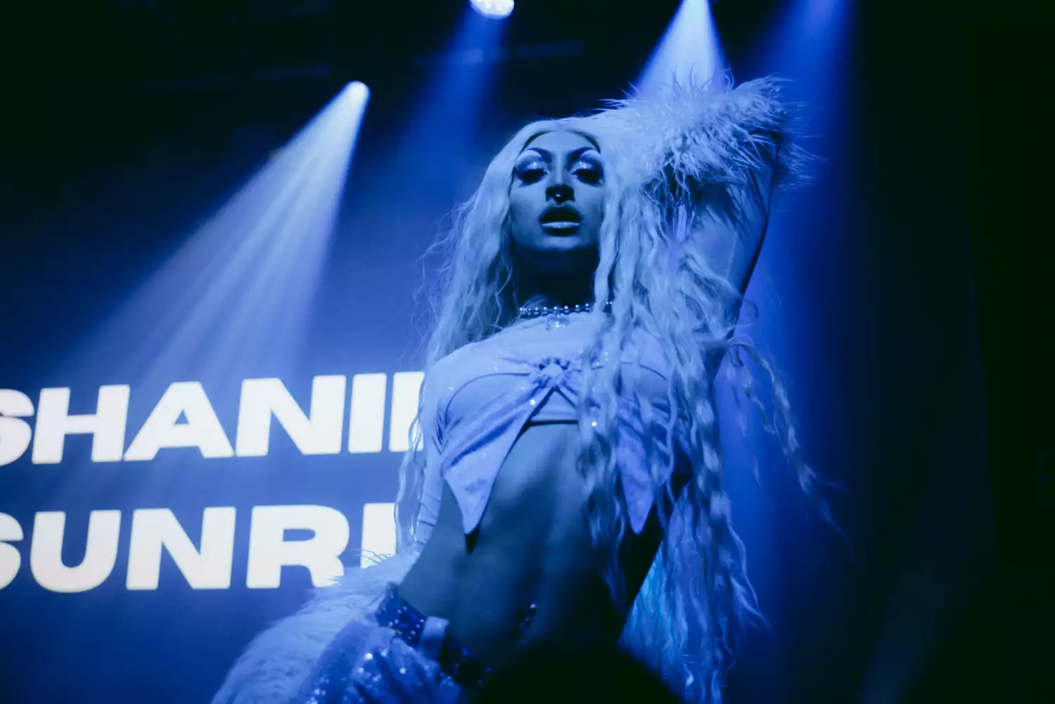 Shanika Sunrise wearing a crop top and posing on stage, with their name in lights behind them