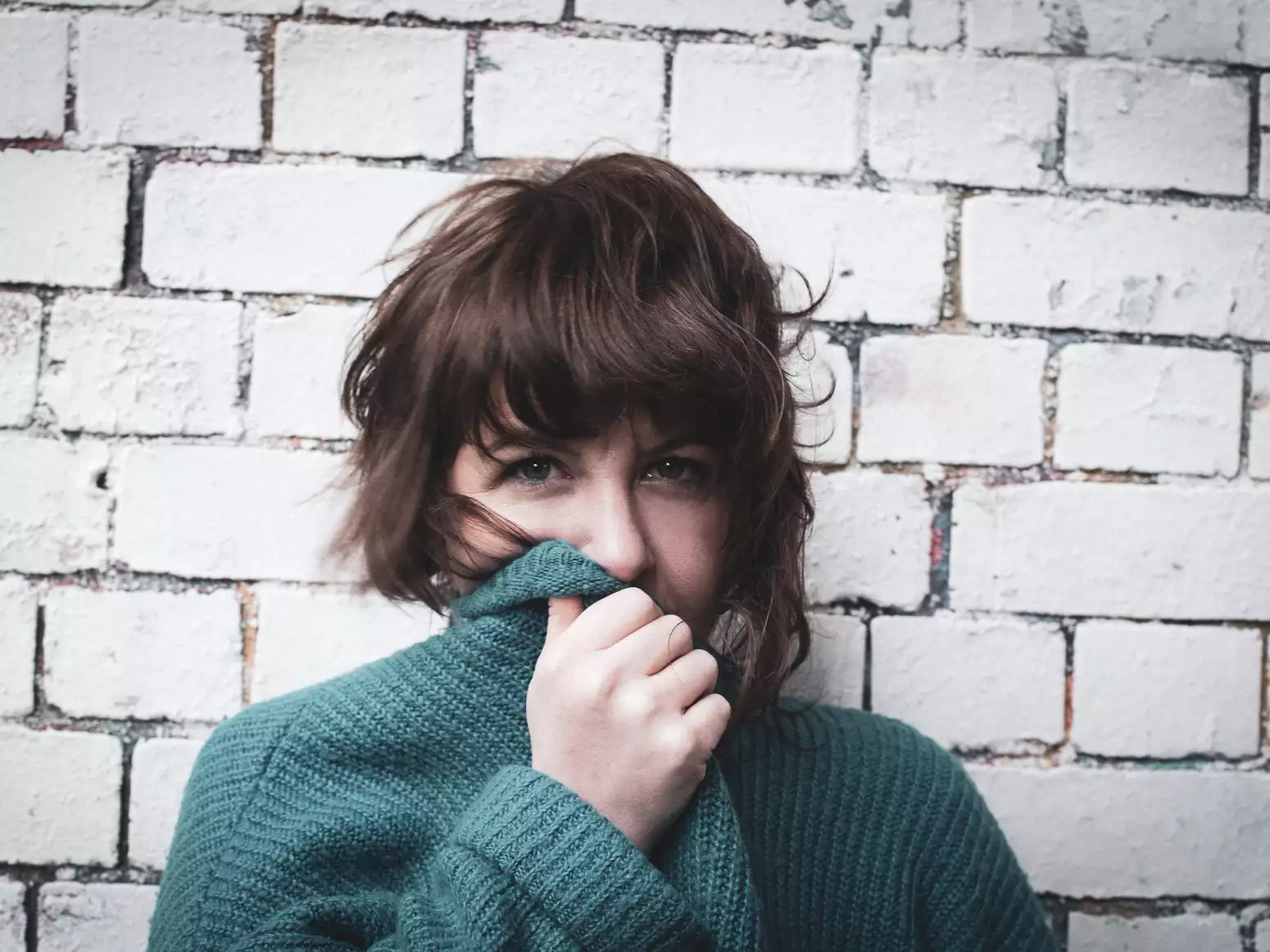 Facing directly into the camera you can see that Test Card Girl is wearing a green cardigan or jumper, she is partially covering her face with the collar of it. She has wavy brown hair and is standing in front of a white brick wall.