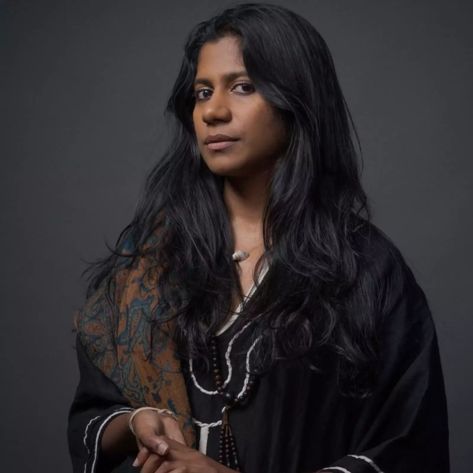 Headshot of ganavya wearing a black shirt and patterned brown scarf on a black background